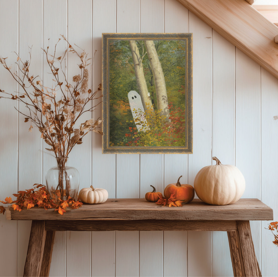 Decorative setup with pumpkins, a vase of dried flowers, and a ghostly painting on a wooden table against a white paneled wall.