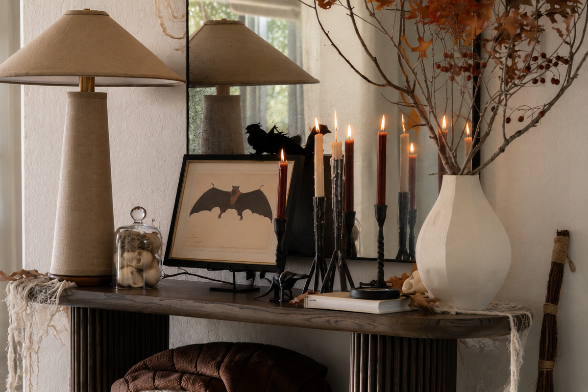 A styled interior console table scene with two beige table lamps, black candlesticks with lit taper candles, a framed bat silhouette print, a white vase with dried branches, and a mirror reflecting the lamps.